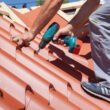 Worker on a roof with electric drill installing red metal tile on wooden house