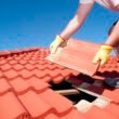 Roof repair, worker with yellow gloves replacing red tiles or shingles on house with blue sky as background and copy space.