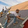 A team of men install watreproofing material and shingles to create a new roof on a rural building.